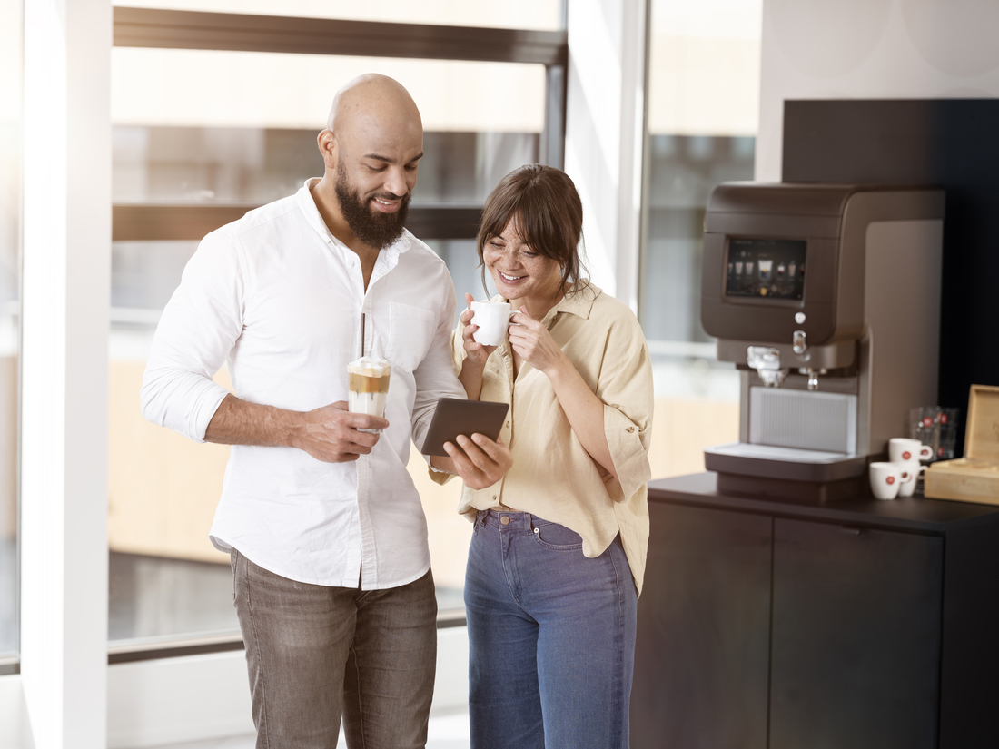 Twee mensen overleggen bij koffiemachine met tablet en drankjes in lichte kantooromgeving.