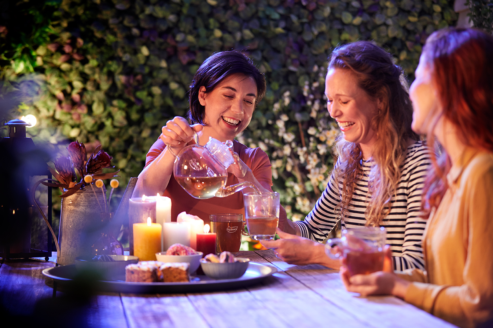 Drie vrouwen zitten gezellig samen aan een houten tafel buiten in de avond, omringd door brandende kaarsen en bloemen. Eén vrouw schenkt thee uit een glazen theepot in een glas dat een andere vrouw vasthoudt